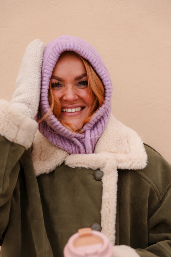 Nainen hymyilee katsoen kameraan päällään laventelin sävyinen huppu ja lämmin takki.